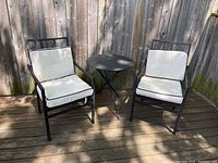 Two black metal patio chairs with cream cushions and black piping seated on wooden deck and a round black metal side table between them.