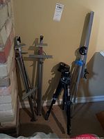 Photo showing a Slik Universal U212 Deluxe tripod standing next to three silver metal portable easels, all placed against a wall with beige carpeted floor.