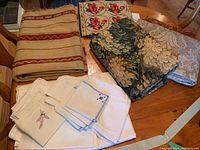 Photo of folded vintage table linens including different types of tablecloths, napkins, printed fabric, and embroidered tea towels on a wooden table.