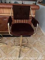 Single brown velvet bar stool with high back, armrests, metal swivel base and footrest. Seen on tiled floor in front of brown leather sofa.