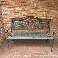 Steel garden bench placed against a brick wall, showing front and side armrests, ornamental backrest, and perforated seat surface.