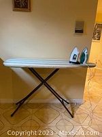 Ironing board set up against a wall, showing the full length of the board and the two irons resting on top.