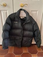Black Columbia winter jacket hanging on a golden door handle, size Large visible on inner tag.