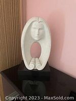 Front view of white plaster sculpture showing stylized woman's face with hollow center and hair flowing down on a black base