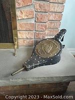 Black vintage fireplace blower with a brass relief medallion of a bearded man wearing a hat and smoking a pipe.