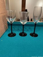 Three glasses displayed side by side on a table, shows three different styles of glasses with black stems: a standard rounded wine glass, a square/bulbous shaped wine glass, and a tall champagne flute.