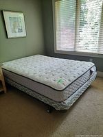 Bed with mattress and box spring on metal frame next to window and nightstand.