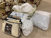 Photo showing various bedding items including pillows, comforters, and blankets on carpet with floral furniture in background.