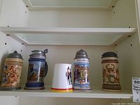 Five beer steins displayed on a white shelf, four have pewter lids, varying in size and design.