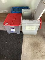 Shows plastic storage bins with red lids and one blue lid, some lids removed, bins stacked and on floor.