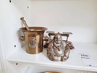 Photo showing the full lot including brass mortar with pestle, brass Buddha statue, and brass decorative scale with weights on a white shelf.