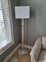 Full view of modern floor lamp standing next to window and couch, showing metal base and white fabric shade.
