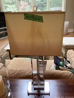 Front-facing image of a modern table lamp with a clear glass base, rectangular beige shade, and finial on top, labeled with dimensions 10x5x21 inches.