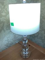 Full view of the turned-on glass table lamp with white cylindrical shade on wooden table against floral wallpaper.