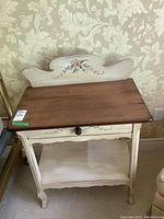Front angle view of hand painted wooden table showing wood top, drawer, bottom shelf and decorative backboard with floral design.
