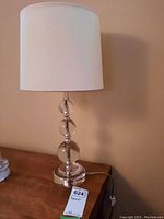 Table lamp with clear glass base and white fabric shade showing some stains on the shade.