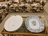 Three large platters placed on a glass and wood table in front of a floral sofa set.