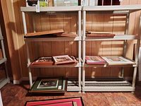 Front view of two shelving units side-by-side with various flattened picture frames and other items on shelves, overall condition visible.