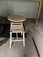 Full view of metal top patio side table with metal base and wood framed stool with woven reed seat beside it in a garage setting.