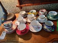 Overall view of 11 tea cups with saucers displaying various floral and decorative patterns in different colors on a wooden table.