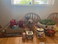 Wide view showing entire lot on wooden table against window with wooden chairs. Includes red pillar candles on metal trays, battery candles, decorative containers, faux plant pot, figurines, and plaque.