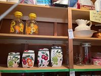 Shelf showing two yellow-lidding glass pitchers with citrus decoration and ceramic canisters with painted fruit including grapes, oranges, and cherries.