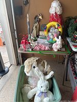 Lot of Easter items displayed on a green table and in a container including bunny stuffed animals, Easter eggs, wreath, and other decorations.