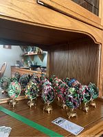Set of 15 Garden Majesty eggs displayed on wooden shelf with floral and butterfly detailed open lattice design, all on gold-tone stands.