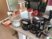 Overview of all kitchen appliances including Rival crock pot, coffee makers, bread maker, lid remover, and electric grill on a table