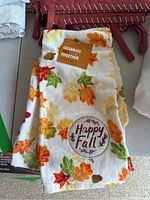 White towel with colorful fall leaves and Happy Fall text folded with rust-colored curtains in background