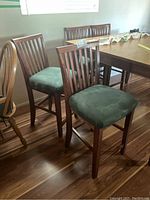 Six wooden pub-style chairs grouped around a table, showing vertical slat backs and green faux suede upholstered seats.
