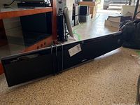 Black Samsung sound bar lying horizontally on the floor, showing front and side profiles with shiny black finish and silver accents.
