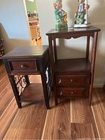 Photo showing two wood side tables side by side on wood flooring. Smaller table has one drawer and decorative metal scrollwork on sides. Taller table has two drawers and an open shelf. Both have dark wood finish.