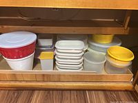 Shelf with assorted Tupperware containers and mixing bowls in various sizes and colors, predominantly white and yellow lids, red and white large mixing bowl.