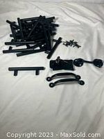 Photo showing scattered black hardware including drawer pulls, door pulls, latch, and hasp on white fabric.