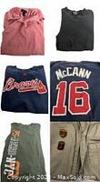 Pile of folded clothing including pink hoodie, dark gray or navy sweatshirt, Atlanta Braves shirt with McCann 16, green graphic long sleeve tee, and khaki pants.