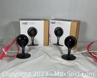Two Nest Security Cameras with black stands, power cords, and original boxes shown from the front on a white surface.
