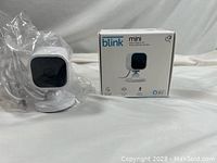 Blink Mini camera in front of its original box, camera wrapped in plastic indicating new condition.