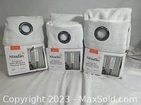 Three new Studio 3B curtain panels in white linen with black metal grommets for hanging. Packaging is visible with product label and description showing dimensions and blackout feature.
