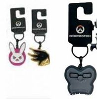 Close-up photo of three Overwatch metal keychains showing D.Va pink bunny logo, Mercy gold wing logo, and Winston gorilla face. Keychains are attached to black branded hooks.