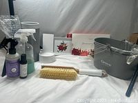 Full display of all cleaning supply set components arranged on white cloth including two metal caddies labeled 'Grove,' a glass spray bottle, liquid bottles for hand sanitizer and air freshener, a natural fiber brush with wooden handle, and a white stoneware tray.
