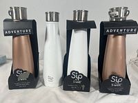 Four Sip by S'well tumblers displayed side by side, two in bronze metallic finish and two in white finish, all with carry caps and branded packaging labels.