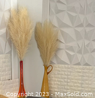 Two sets of faux pampas grass arranged in tall brown ceramic vases, showing beige fluffy plumes against a white textured wall.