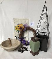 Image of entire indoor/outdoor decor collection showing a concrete bowl, metal trellis pot, decorative green vase, metal candle holder, wreath with felt flowers, Welcome garden flag, garden stake, and metal wall hook.