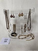 Full view of clear acrylic jewelry stand holding various necklaces, bracelets, and pairs of drop earrings. Anne Klein watch with black strap seated on base of stand. Pearl-tone beaded necklace also on stand base.