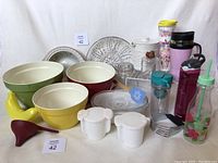 Photo showing kitchen lot including various sized nesting mixing bowls in yellow, green, and red, vintage Tupperware cream and sugar containers, multiple water bottles in different styles and colors, Corningware casserole dish, glass serving platters and bowl, yellow banana keeper, red funnel, and other kitchen items grouped together