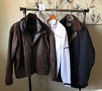 Three men's jackets on rack: brown leather/suede Cruze jacket, white fleece-lined zip-up sweater, navy blue Rainforest coat