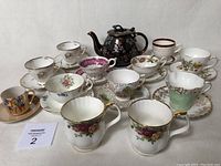 Front view of 10 teacups with saucers and 2 solo teacups with hand-painted dark floral teapot in the back