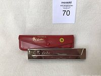 Photo showing the harmonica and its red case with gold branding and 'G' sticker, laid flat on white surface.