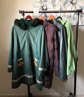 Four women's jackets hanging on rack including green Indigenous ulu coat, brown down puffer, black leather jacket and green windbreaker, showing front views.